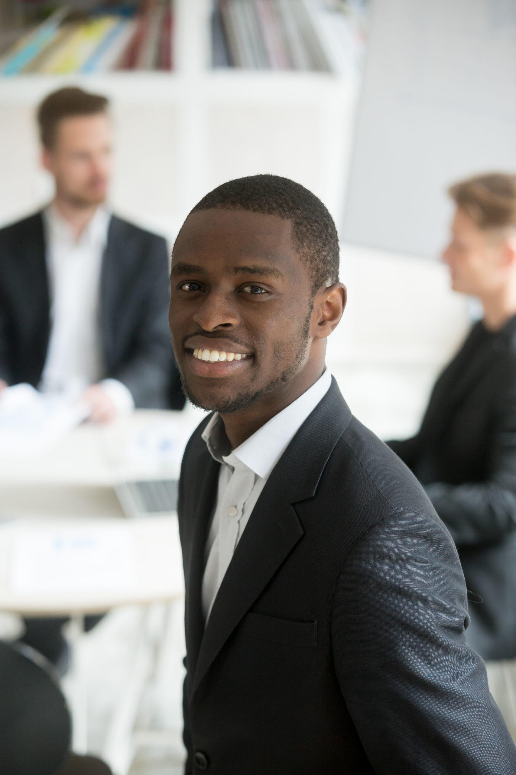 Smiling attractive young african american businessman in suit looking at camera, headshot vertical portrait of black professional executive manager posing with business partners team at background